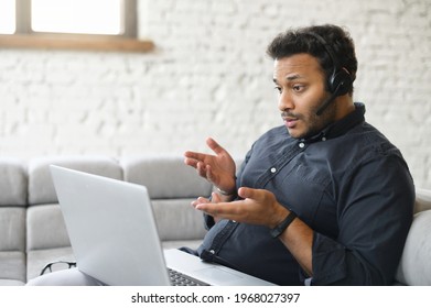 Concentrated Indian Man Wearing Headset Holding Virtual Video Conference Sitting On The Couch At Home With A Laptop Computer On The Laps, Multiethnic Guy Studying Online, Involved Webinar