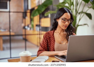 Concentrated latin business woman working on laptop wearing eyeglasses. Focused woman entrepreneur working on laptop in office lobby. Young serious multiethnic businesswoman working online on computer - Powered by Shutterstock