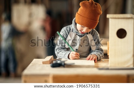 Concentrated little boy drawing sketch on wooden plank while making bird house in craft workshop