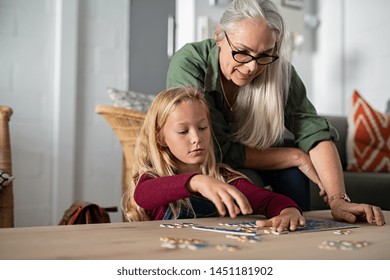 Concentrated Little Girl Doing Jigsaw Puzzle With Old Grandmother At Home. Cute Little Granddaughter Playing Puzzle At The Table With Senior Granny. Smart Child Assembling Pieces With Mature Woman.