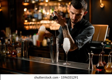Concentrated male bartender pouring a liqour from the jigger to a professional steel shaker on the bar counter