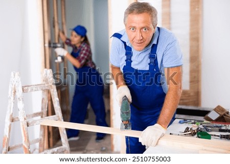 Concentrated male builder in uniform using a jigsaw machine on wooden plank at the apartment