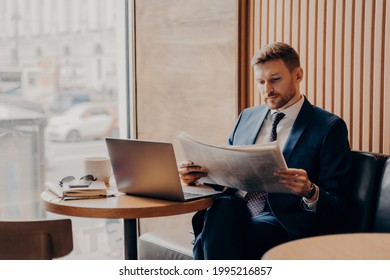 Concentrated Male Business Company Worker In Blue Formal Suit Sitting In Corner Of Cafe Next To Big Window, Reading Newspaper Closely, Open Work Laptop And Coffee In Front Of Him On Table