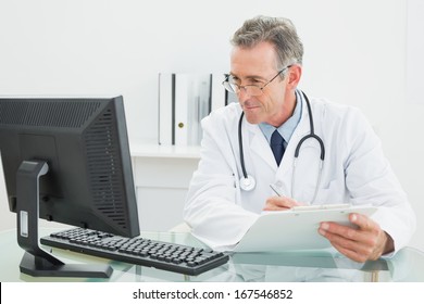 Concentrated Male Doctor With Report Looking At Computer Monitor At Desk In Medical Office