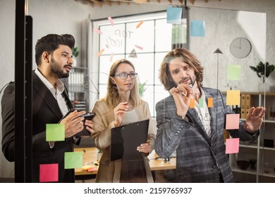 Concentrated Male And Female Entrepreneurs With Clipboard And Digital Tablet Writing Notes On Glass Wall At Modern Office. Group Of Multiethnic Business People Having Brainstorm Session In Boardroom.