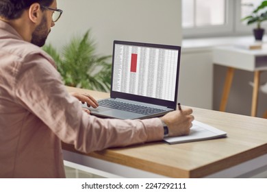 Concentrated male financier using laptop working in office with data entered in spreadsheet. Man fills out paper documents and enters data into electronic files marked in red on laptop screen.
