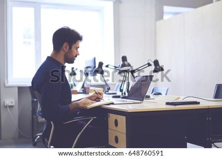 Concentrated male journalist browsing information from social networks preparing article noting ideas, professional handsome copywriter working on creation advertising content for marketing website  