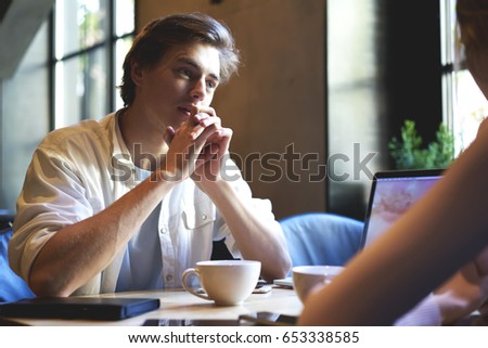 Concentrated male journalist listening carefully interlocutor during friendly conversation in cafeteria.Serious young hipster guy communicating with employer about work in coffee shop indoors
