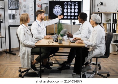 Concentrated Male Medical Specialist Pointing On Screen With Brain Scan And Discussing Diagnosis With Diverse Colleagues. Multiracial Team Thinking About Ways Of Successful Treatment.