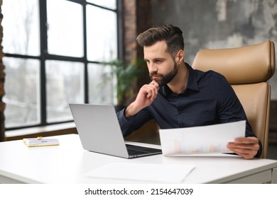 Concentrated Male Small Business Owner Holding Papers And Looking At The Laptop Screen While Sitting In Modern Loft Office. Successful Contemporary Man Manager In Casual Wear Looking At The Document