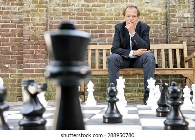 Concentrated Man, Thinking Strategically About His Next Move, Sitting On A Wooden Bench In Front Of A Brick Wall During An Outdoor Chess Game Using Life Sized Chess Pieces And Chess Board