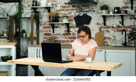 Concentrated Mature Woman Taking Notes While Using Laptop In The Kitchen