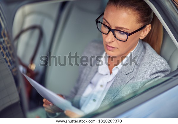 Concentrated middle aged businesswoman wearing\
eyeglasses reading documents while sitting on back seat in the car,\
preparing for a\
meeting