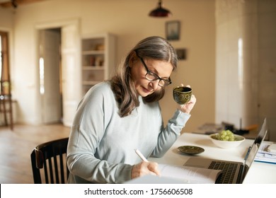 Concentrated Middle Aged Female Teacher In Glasses Sitting At Desk Drinking Coffee, Handwriting In Copybook, Making Lesson Plan, Using Portable Computer, Teaching Online From Home. Age And Technology