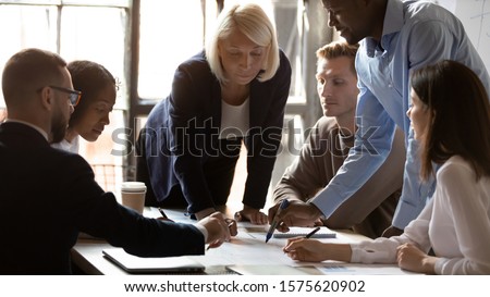 Concentrated middle aged female team leader leaning over table, looking at african american young employee pointing at financial document. Group of international specialists discussing project details