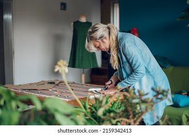 Concentrated Middle Aged Woman Tailor Cuts With Scissors Pattern Dress Detail On Sketch Lines For Sewing Machine. Workplace Of A Dressmaker. Handmade Concept. Small Business Owner.