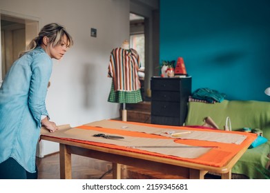 Concentrated Middle Aged Woman Tailor Cuts With Scissors Pattern Dress Detail On Sketch Lines For Sewing Machine. Workplace Of A Dressmaker. Handmade Concept.