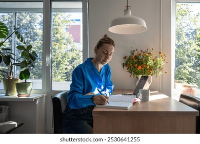 Concentrated middle aged woman works at home, planning day jotting down diary, comfortable sitting at table with laptop surround by houseplants. Focused female writing notes in day planner in morning - Powered by Shutterstock