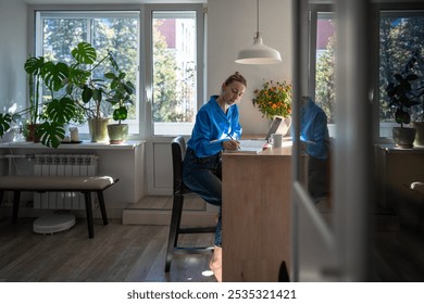 Concentrated middle aged woman works at home, planning day jotting down diary, comfortable sitting at table with laptop surround by houseplants. Focused female writing notes in day planner in morning