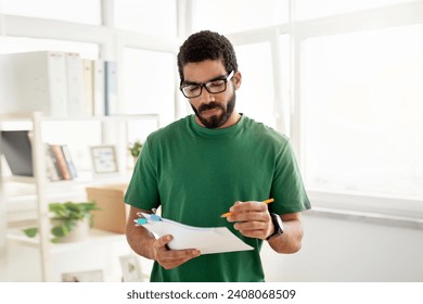 Concentrated middle eastern man with glasses and a beard, wearing a green t-shirt, reads from a paper document while holding a pen, standing in a bright office environment - Powered by Shutterstock