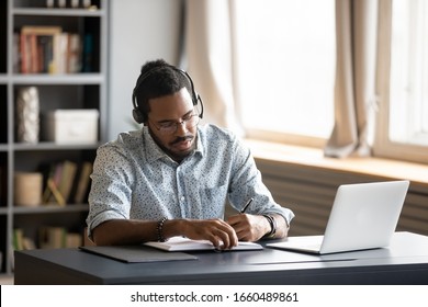 Concentrated millennial african american guy wearing earphones, listening to favorite music while planning workday. Focused young biracial businessman watching educational lecture. writing down notes.
