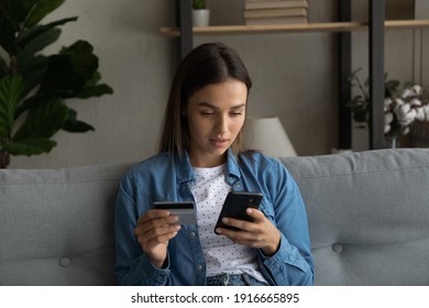 Concentrated millennial female sit on sofa at living room hold plastic credit card provide cashless payment transaction via electronic bank. Focused young lady check card account balance at phone app