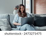 Concentrated millennial girl sit on couch working on laptop browsing internet at home during lazy weekend, focused young woman freelancer busy using computer surfing wireless web shopping online