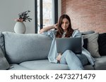 Concentrated millennial girl sit on couch working on laptop browsing internet at home during lazy weekend, focused young woman freelancer busy using computer surfing wireless web shopping online