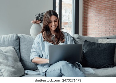 Concentrated millennial girl sit on couch working on laptop browsing internet at home during lazy weekend, focused young woman freelancer busy using computer surfing wireless web shopping online