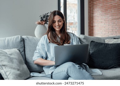 Concentrated millennial girl sit on couch working on laptop browsing internet at home during lazy weekend, focused young woman freelancer busy using computer surfing wireless web shopping online