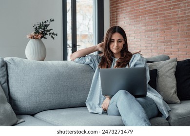 Concentrated millennial girl sit on couch working on laptop browsing internet at home during lazy weekend, focused young woman freelancer busy using computer surfing wireless web shopping online