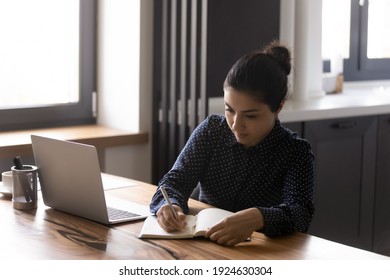 Concentrated Millennial Indian Woman Sit At Desk At Home Make Notes Study Distant On Laptop. Focused Young Ethnic Female Employee Watch Webinar Take Online Course Or Work On Internet On Computer.