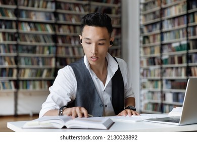 Concentrated millennial male Korean student in earbuds listening favorite lounge music or educational lecture while reading book, preparing for exam or doing research alone in college library. - Shutterstock ID 2130028892