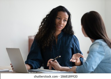 Concentrated multiethnic female employees sit at desk in office work together using computer. Focused women colleagues coworkers cooperate on laptop, brainstorm discuss business ideas at meeting.