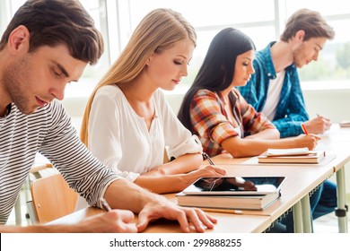 Concentrated On Exam. Group Of Young Students Writing Something In Their Note Pads While Sitting In A Row At Their Desks