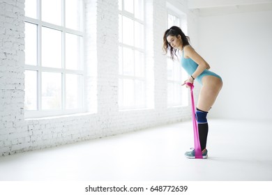 Concentrated On Targets Female Using Elastic Band For Doing Stretching Exercise Tensing Butt Muscles,slim Sportswoman Keeping Fit Healthy Lifestyle In White Interior Gym On Promotional Background