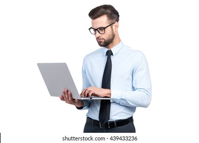 Concentrated on work. Confident young handsome man in shirt and tie working on laptop while standing against white background 