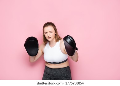 concentrated overweight girl exercising with boxing pads while looking at camera on pink