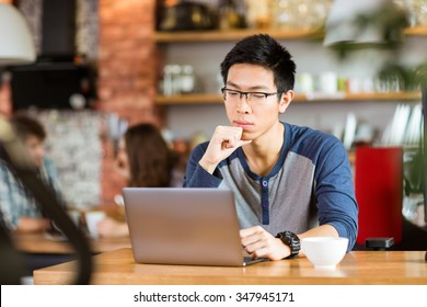 Concentrated pensive young handsome asian man in glasses sitting in cafe, thinking and using laptop