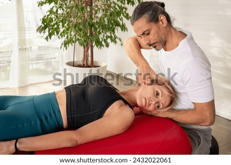 Concentrated physiotherapist manipulating spine of patient to alleviate neck pain in alternative medicine during consultation in modern clinic with plants and glass wall