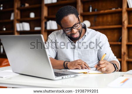Concentrated positive hispanic man in eyeglasses thinking, taking notes while sitting at table with laptop and smartphone at home, author writing an idea for his book, study online, e-learning concept