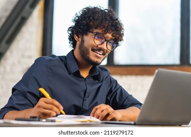 Concentrated Positive Indian Man In Eyeglasses Thinking, Taking Notes While Sitting At The Table With Laptop And Smartphone At Home. Author Writing Idea For His Book, Study Online. E-learning Concept