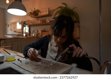 Concentrated precise craftswoman with gas burner sitting at workbench and making handmade accessories in workshop - Powered by Shutterstock