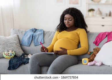 Concentrated Pregnant Black Woman Sitting On Sofa At Home With Notebook And Pen, Making To Do List Before Delivery, Free Space. Pretty African American Single Mother Getting Ready For Baby Coming