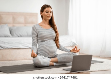 Concentrated pregnant woman seated on a yoga mat, using a laptop for online prenatal classes or relaxation - Powered by Shutterstock