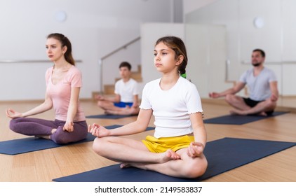 Concentrated Preteen Girl Sitting On Mat In Lotus Position During Yoga Class With Family In Fitness Center..