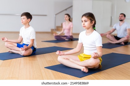 Concentrated Preteen Girl Sitting On Mat In Lotus Position During Yoga Class With Family In Fitness Center..