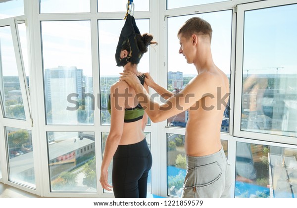 Concentrated qualified\
young male yoga trainer with naked torso correcting shoulders of\
woman with neck inside of cervical traction device, he assisting\
her to straighten\
core