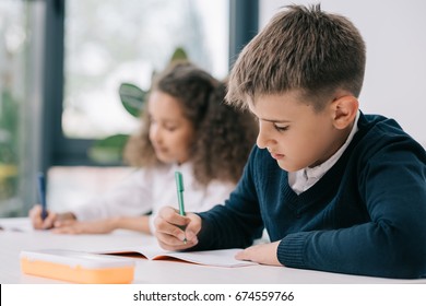 Concentrated schoolboy sitting at desk and writing in exercise book with classmate sitting behind - Powered by Shutterstock