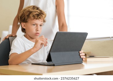 Concentrated schoolkid browsing tablet with stylus while studying in classroom with crop faceless teacher - Powered by Shutterstock